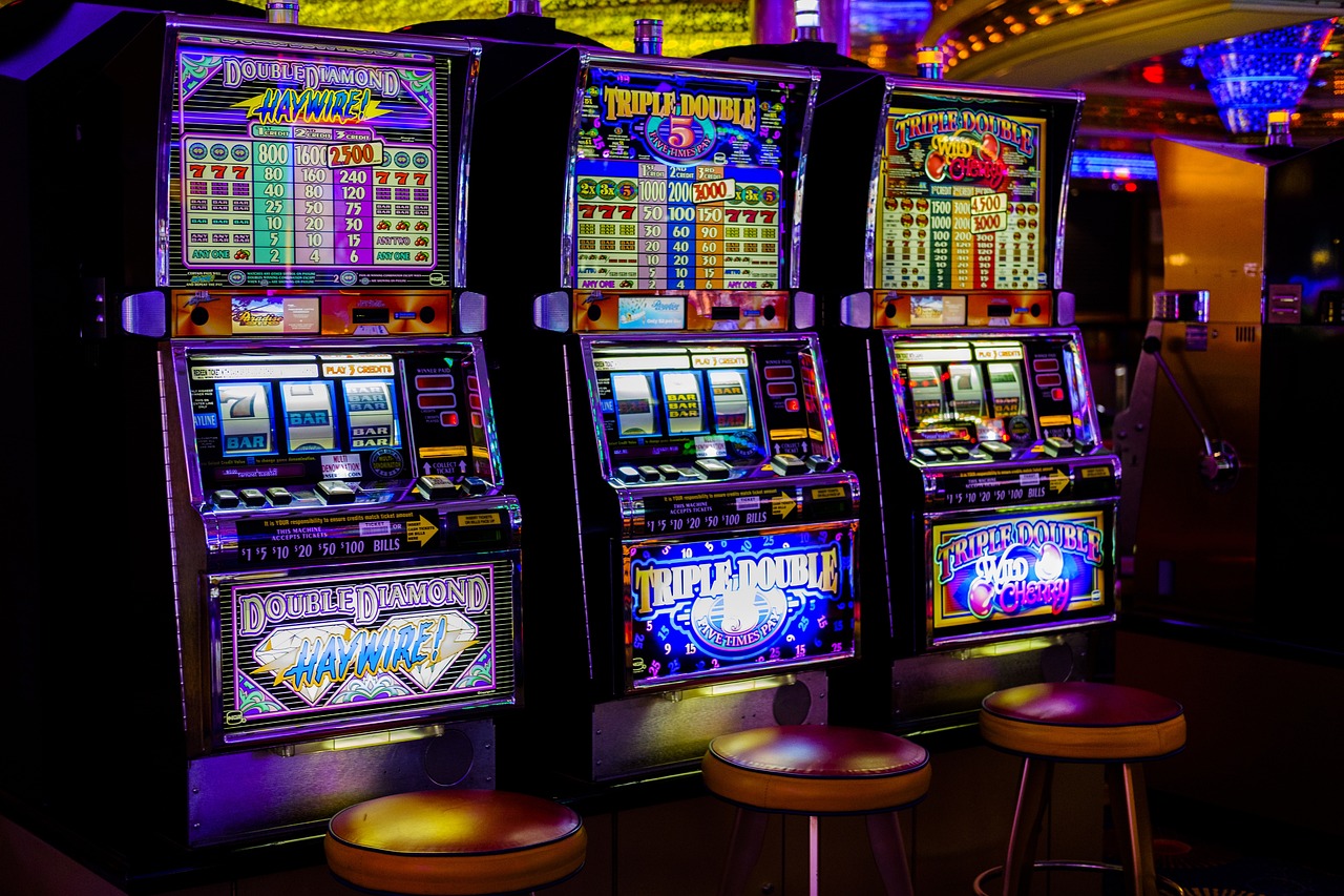 Auf dem Bild befinden sich drei Spielautomaten nebeneinander. Vor den Slot-Maschinen steht jeweils ein Hocker. 