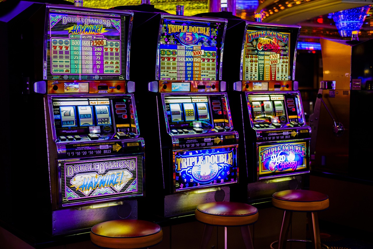 Auf dem Bild befinden sich drei Spielautomaten nebeneinander. Vor den Slot-Maschinen steht jeweils ein Hocker.